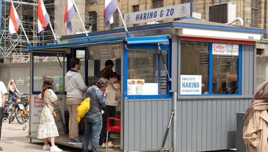 The photo was taken at Nieuwezijds Voorburgwal 200 in Amsterdam-Centrum, behind Dam Square, and shows herring stall Haring & Zo. It captures a few people waiting to be served. The top of the stall is decorated with Dutch flags.