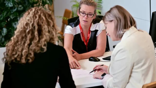 Two women sitting at the desk at the INAmsterdam offices, seen from the back, one of them signing a paper. Employee seen from the front.