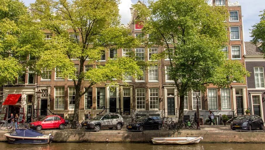 Canal houses seen behind trees with cars and boats parked.